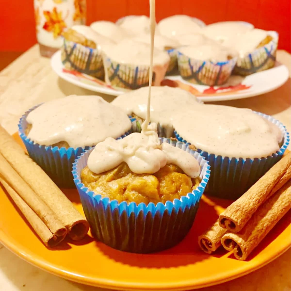 pumpkin muffin in a blue cupcake paper with thick eggnog being poured over it on an orange plate with 3 other muffins and cinnamon sticks