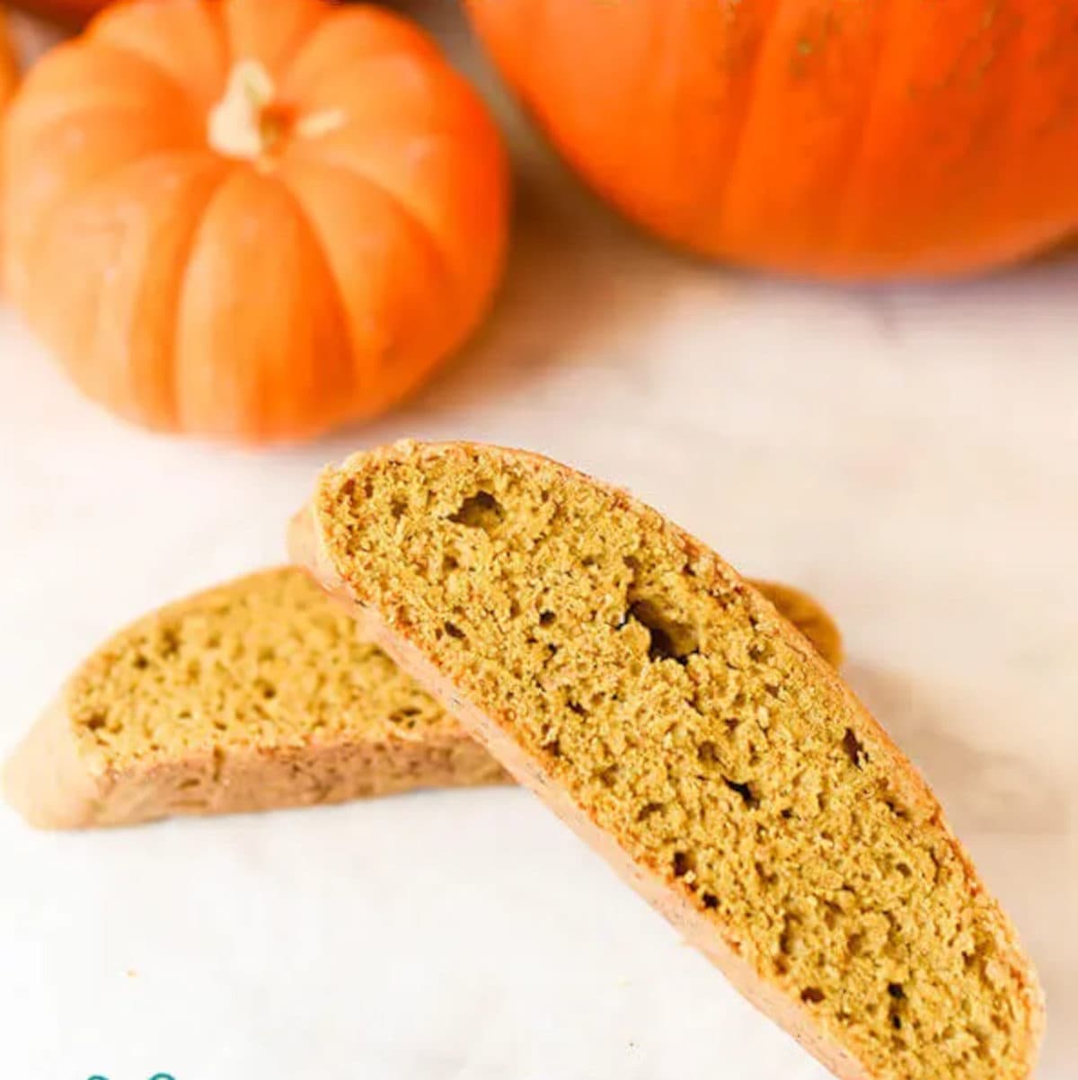 two slices of pumpkin biscotti cookies laying on a white surface beside two orange pumpkins