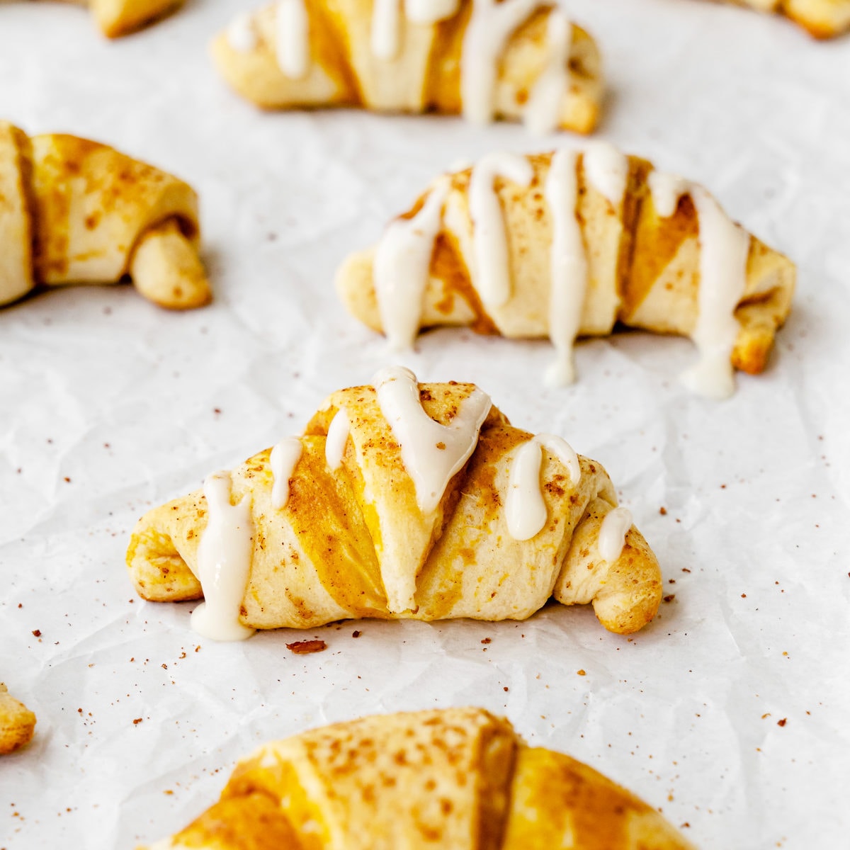 Pumpkin Cream Cheese Crescent Rolls stuffed with pumpkin cheesecake filling, then drizzled with cream cheese frosting on top of white parchment paper.
