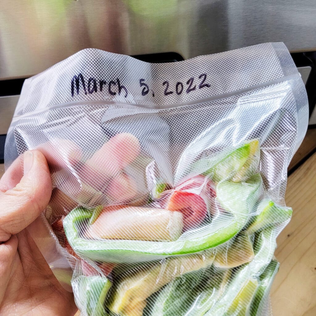 Hand holding a clear vacuum sealed bag of mixed green, red, and yellow bell peppers inside.